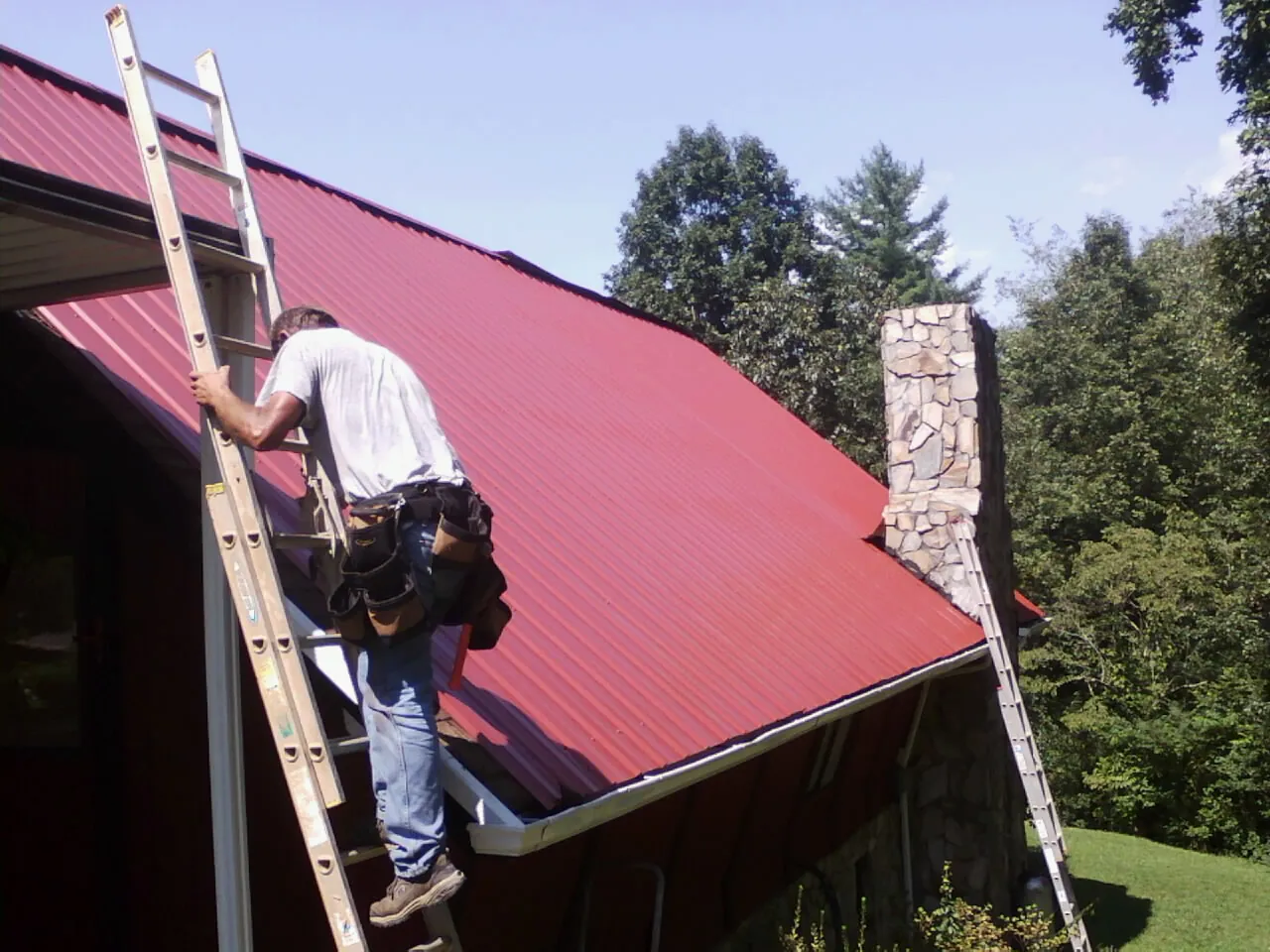 Roofer on a ladder beside a red metal roof and stone chimney — Soffit & Fascia Repair by Roofing Brothers in Homer City